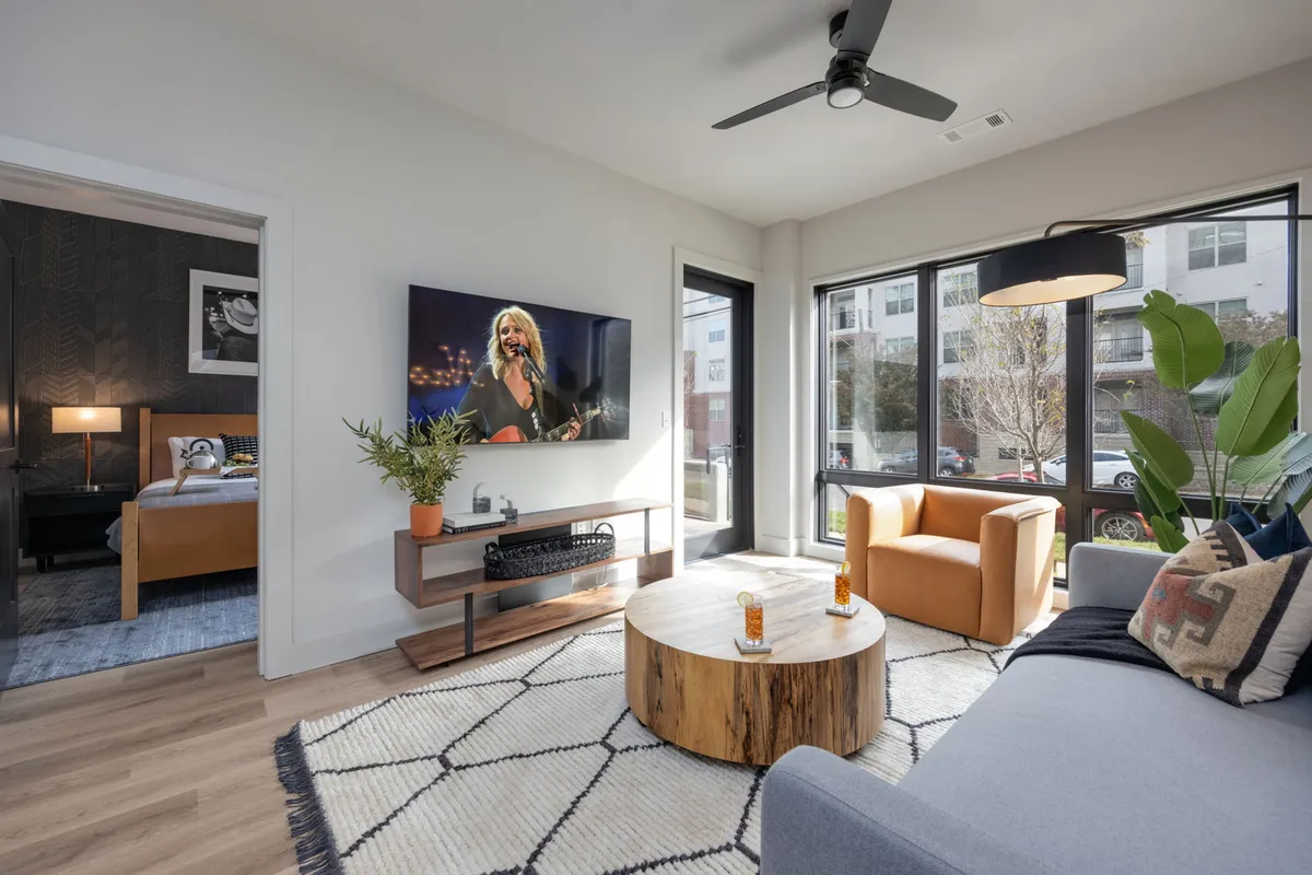 A bright, inviting living room with plush, comfortable seating faces a large TV displaying a musician, reflecting Nashville's music scene. Sunlight pours through expansive windows, illuminating a stylish wooden coffee table and creating a cozy, relaxed ambiance perfect for gatherings or unwinding after a day in Midtown.