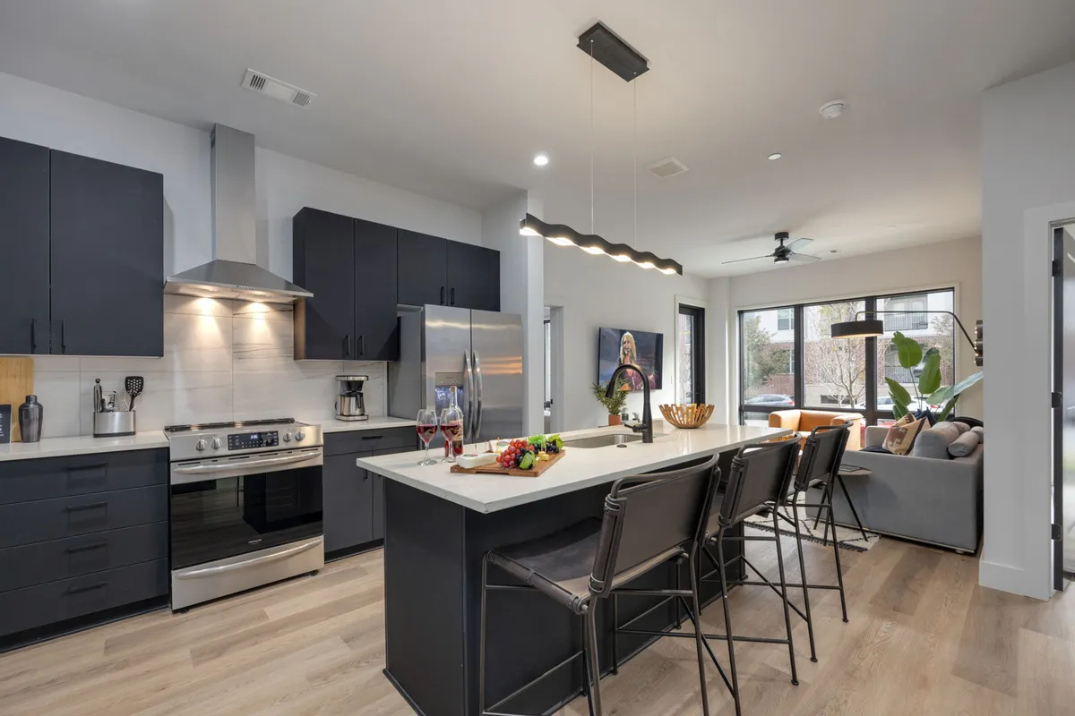 This image features a modern kitchen with a central island, stainless steel appliances, bar stools for seating, and abundant natural light streaming through the windows. A decorative fruit bowl adds a touch of color and homeliness, while the open layout invites both cooking and casual gathering.