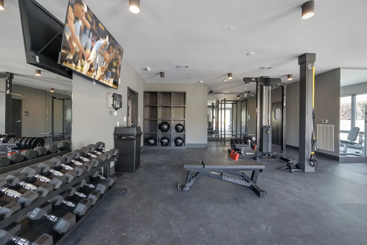 A well-equipped fitness center with a dumbbell rack, weight bench, cable machine, and a mounted television. The layout emphasizes a bright, energetic environment designed for various types of workouts and group fitness activities.