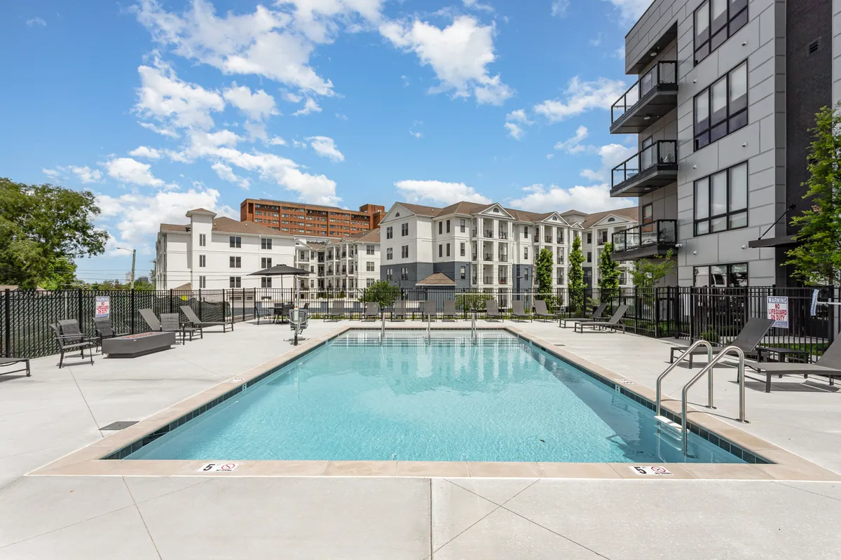 A sparkling swimming pool surrounded by modern apartment buildings, with lounge chairs arranged around the water. The area is illuminated by a warm sunset glow, creating an inviting atmosphere for relaxation, social gatherings, and community events.
